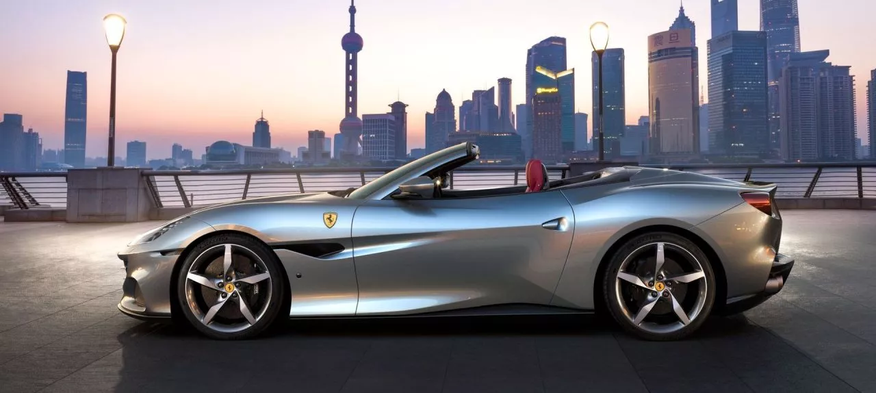 Vista lateral del Ferrari Portofino M con capota bajada frente a un paisaje urbano.