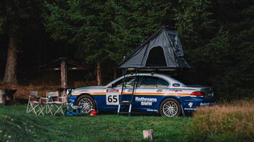 Vista lateral de un BMW Serie 7 con tienda de campaña en el techo, ambientado en una acampada.