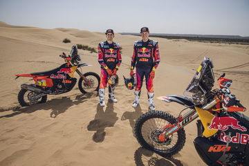 Tres motos de rally KTM con pilotos equipados en el desierto, mostrando sus colores del equipo.