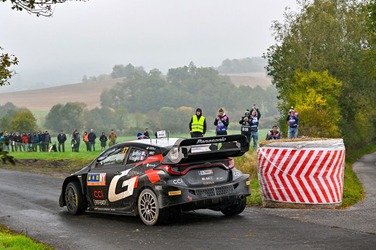 El vehículo de rally mostrando derrape controlado en curva cerrada, público expectante.