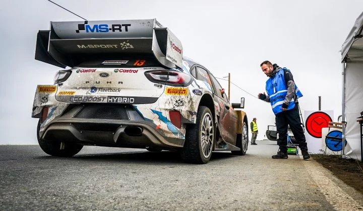 Vista trasera del coche de rally con detalles de su construcción y un equipo técnico al fondo.